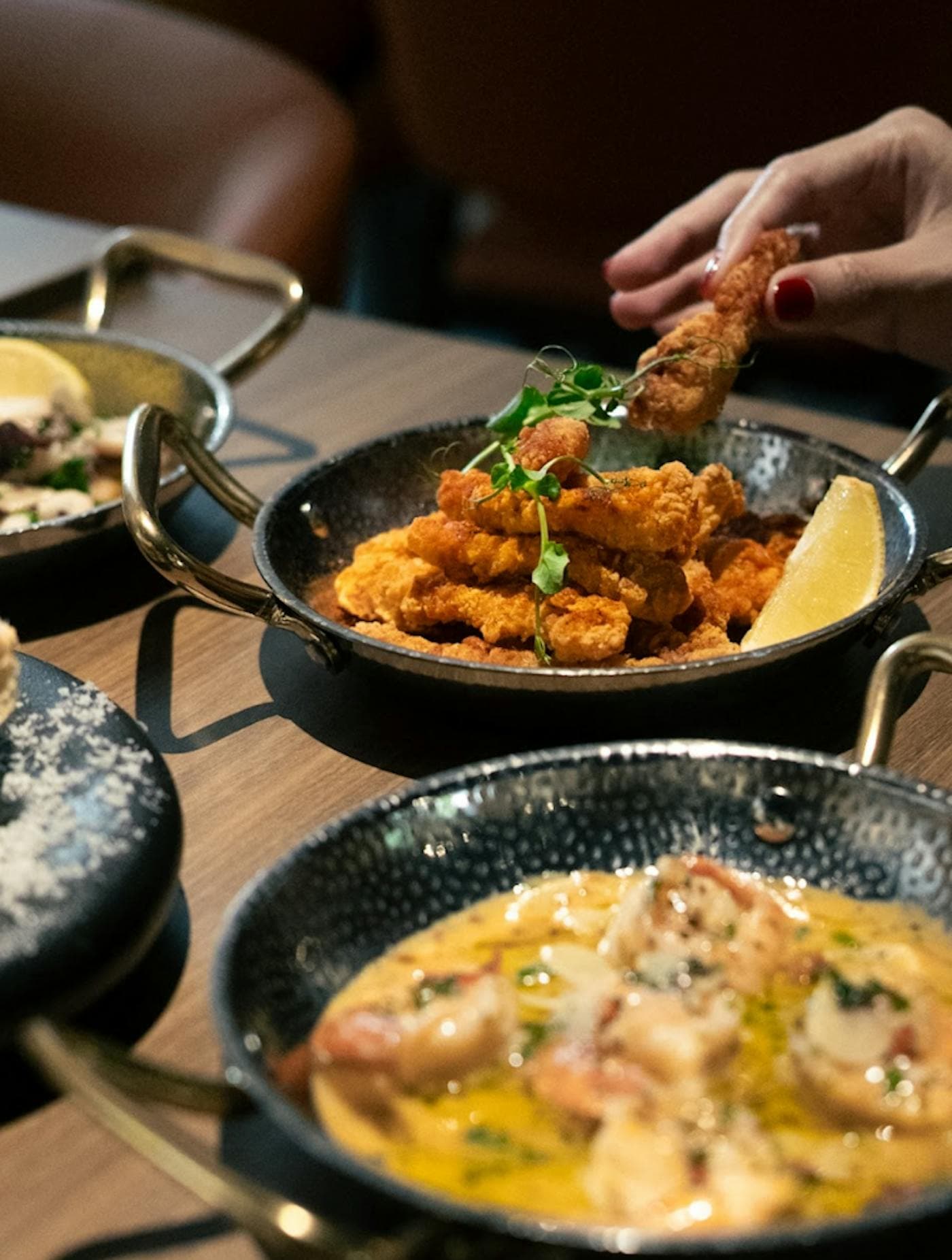 A hand lifting a piece of fried calamari from a black pan, a creamy seafood dish beside it