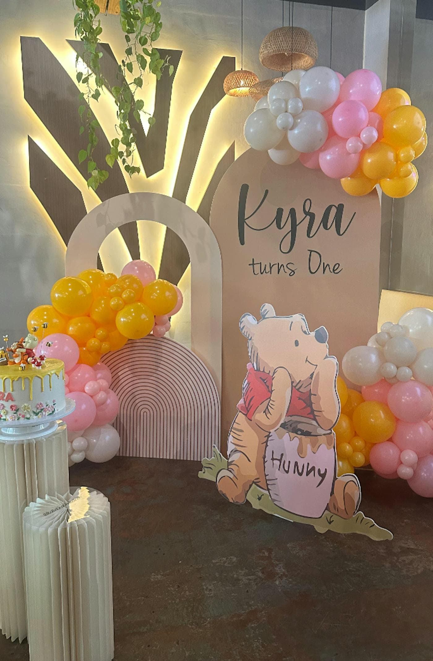 A Kyra Turns One balloon backdrop with a Winnie-the-Pooh cutout and a cake