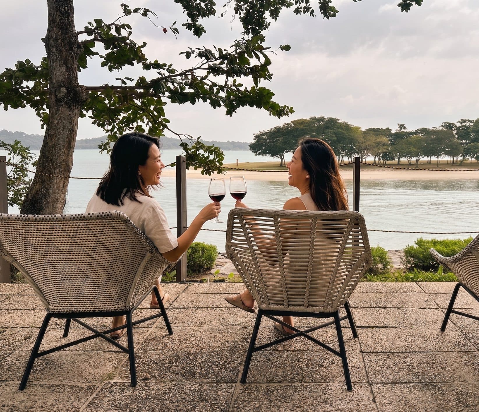 Two friends raising glasses of wine in rope chairs at Canopy Changi Village, the sand and the water behind them