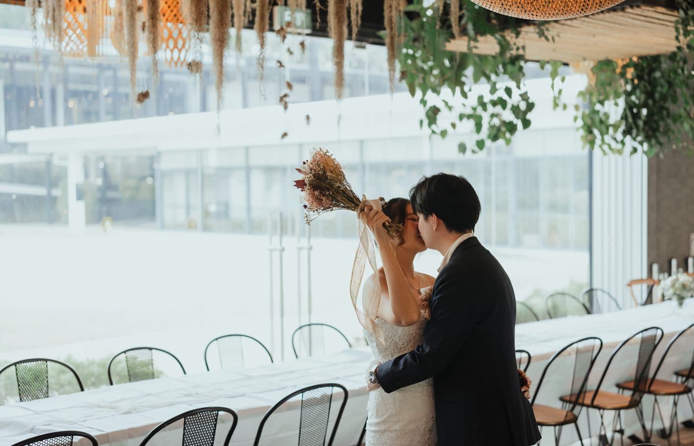 A couple kissing behind their bouquet at a long banquet table under dried florals at Canopy HortPark