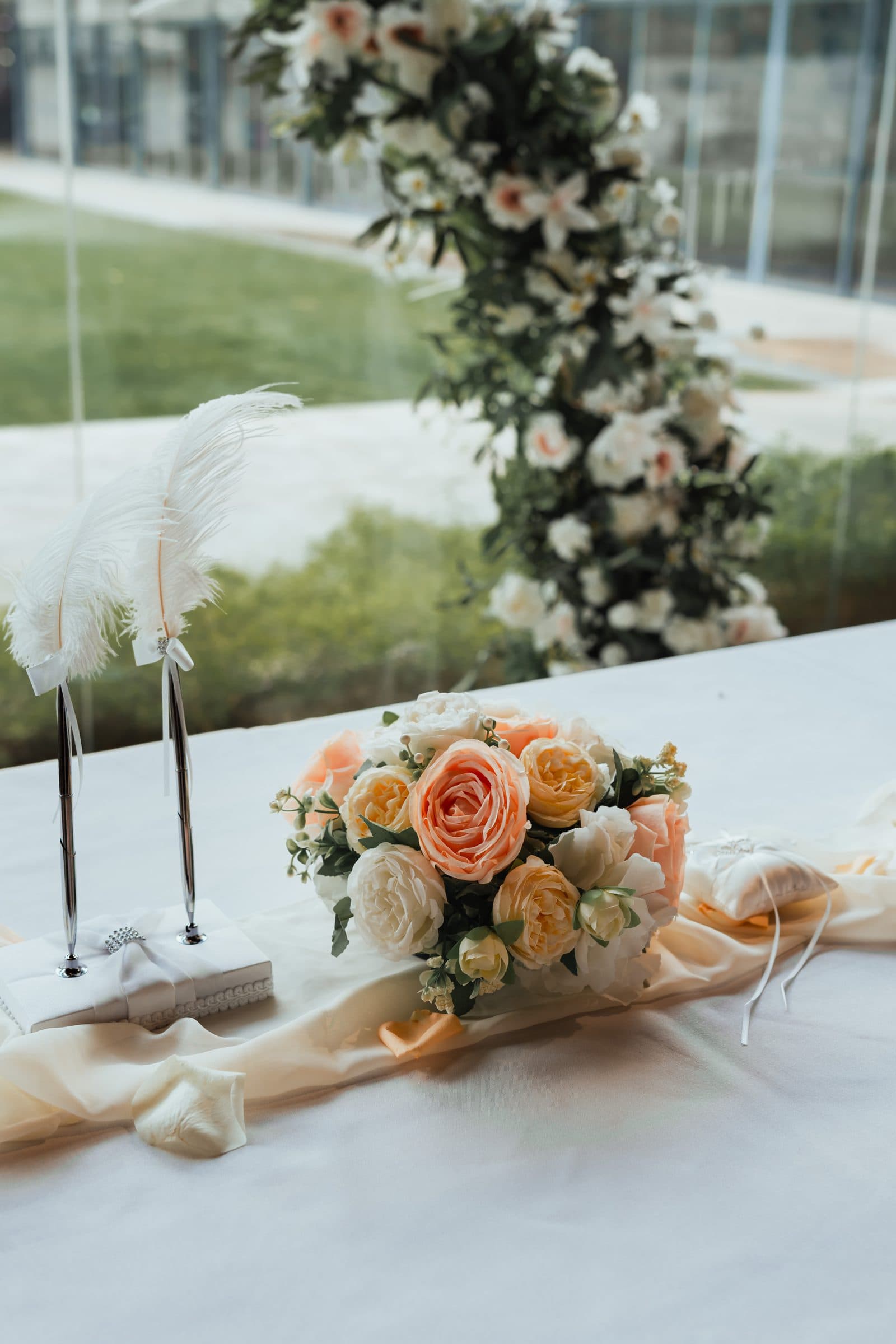 A bouquet and feather pen on the signing table at a Canopy wedding, the floral arch and lawn behind