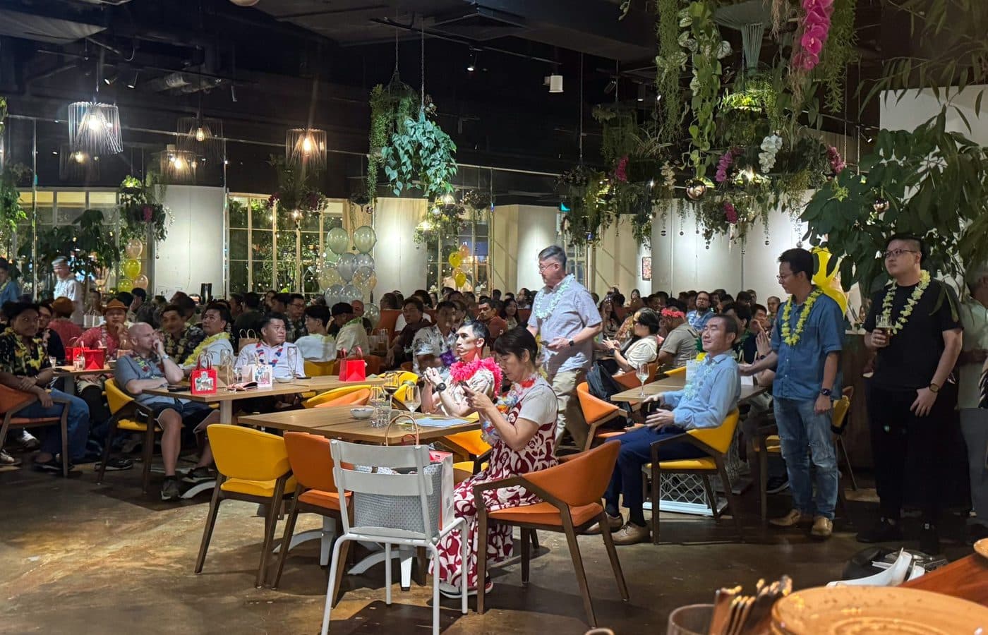 A full room at Canopy Esplanade during a company party, guests in leis under hanging orchids