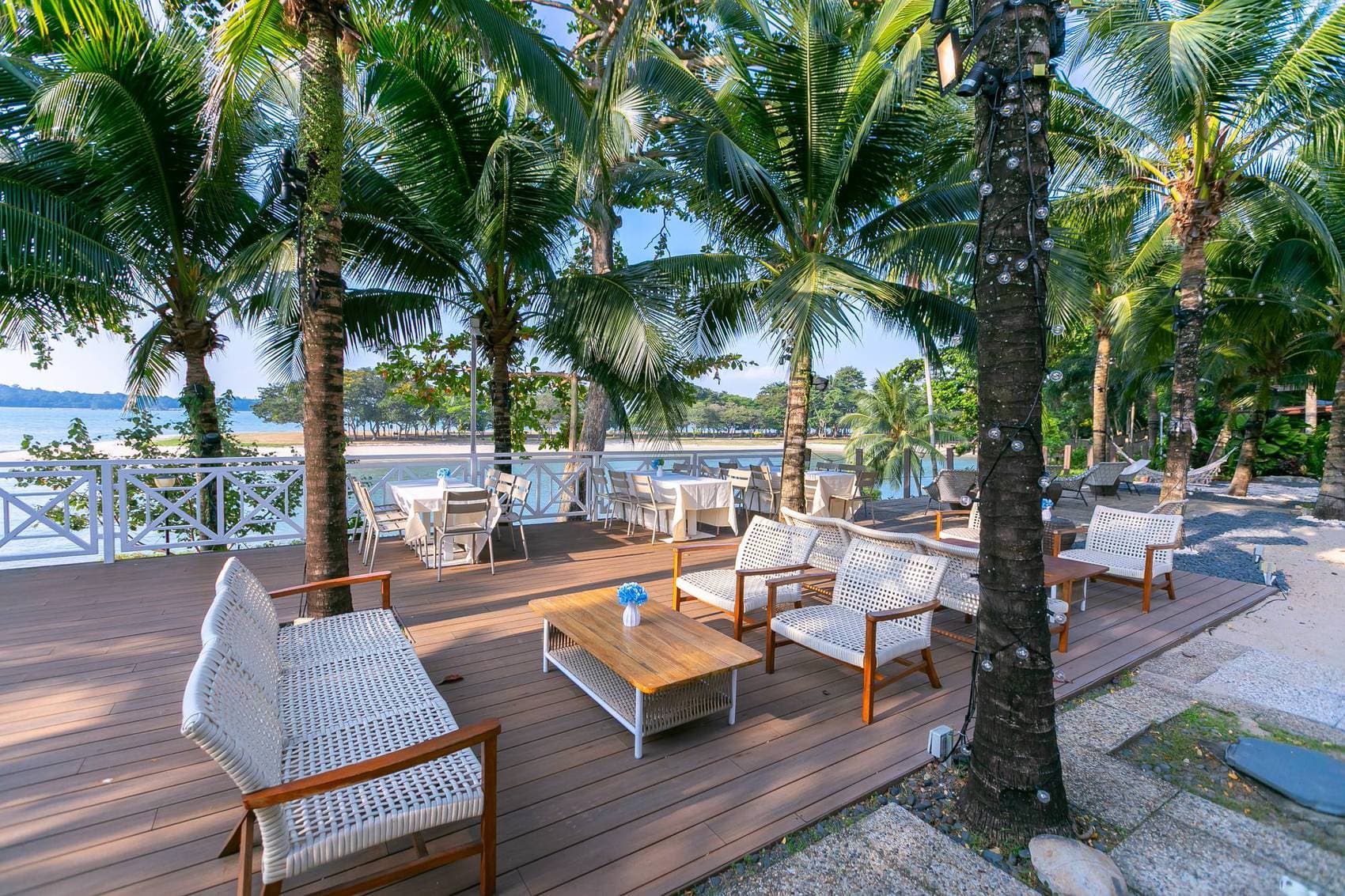 White rope lounge chairs and low timber tables on the deck at Canopy Changi Village, tall palms above and the sea past the railing