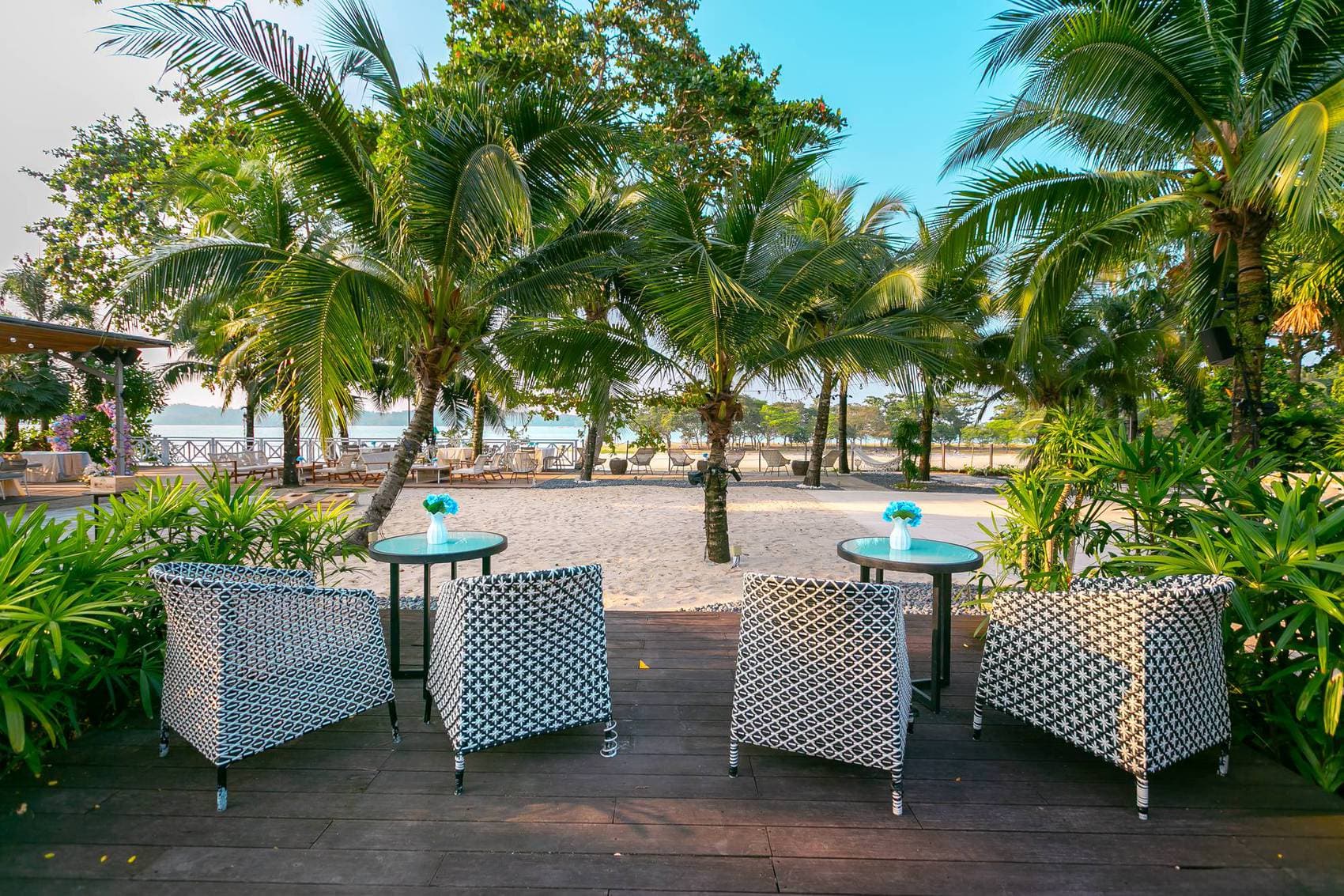 Rattan tub chairs and low tables on the deck at Canopy Changi Village, palms overhead and the sand beyond