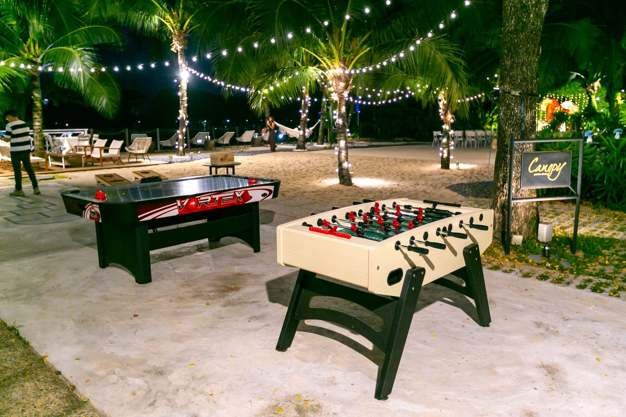 Foosball and air hockey on the sand at Canopy Changi Village at night, festoon lights strung between the palms