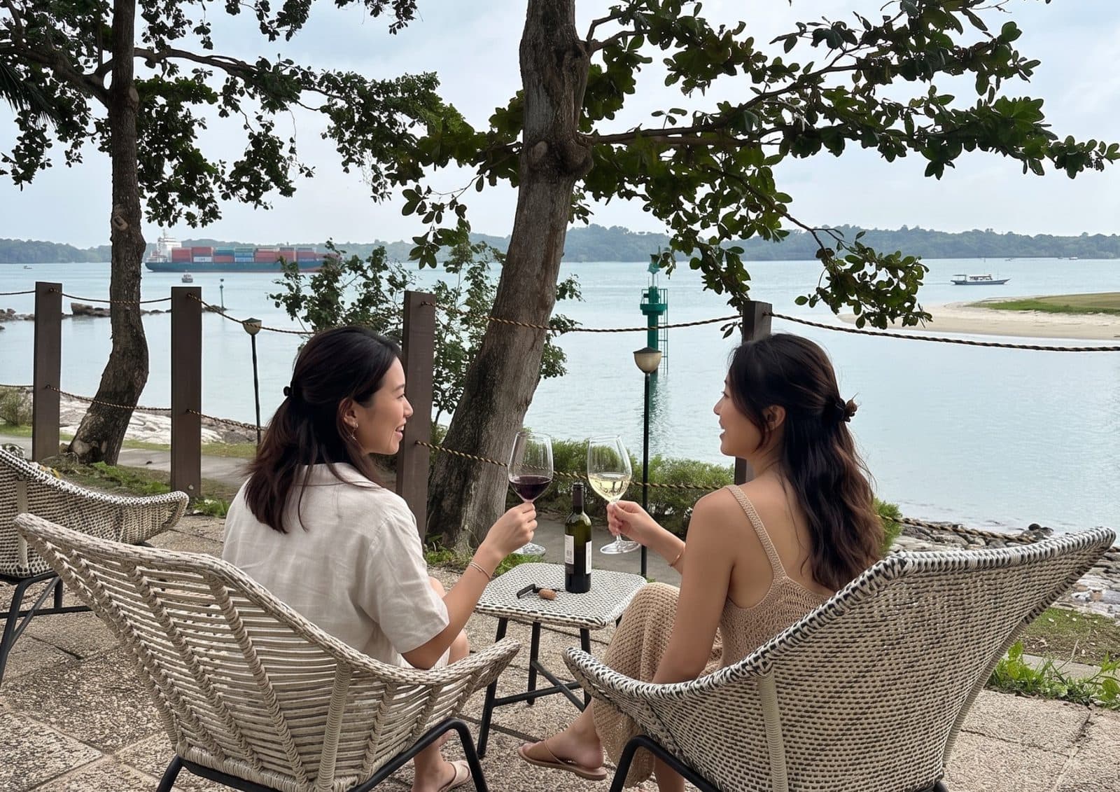 Two friends with wine at a low table at Canopy Changi Village, tree canopy overhead and the shipping lane beyond