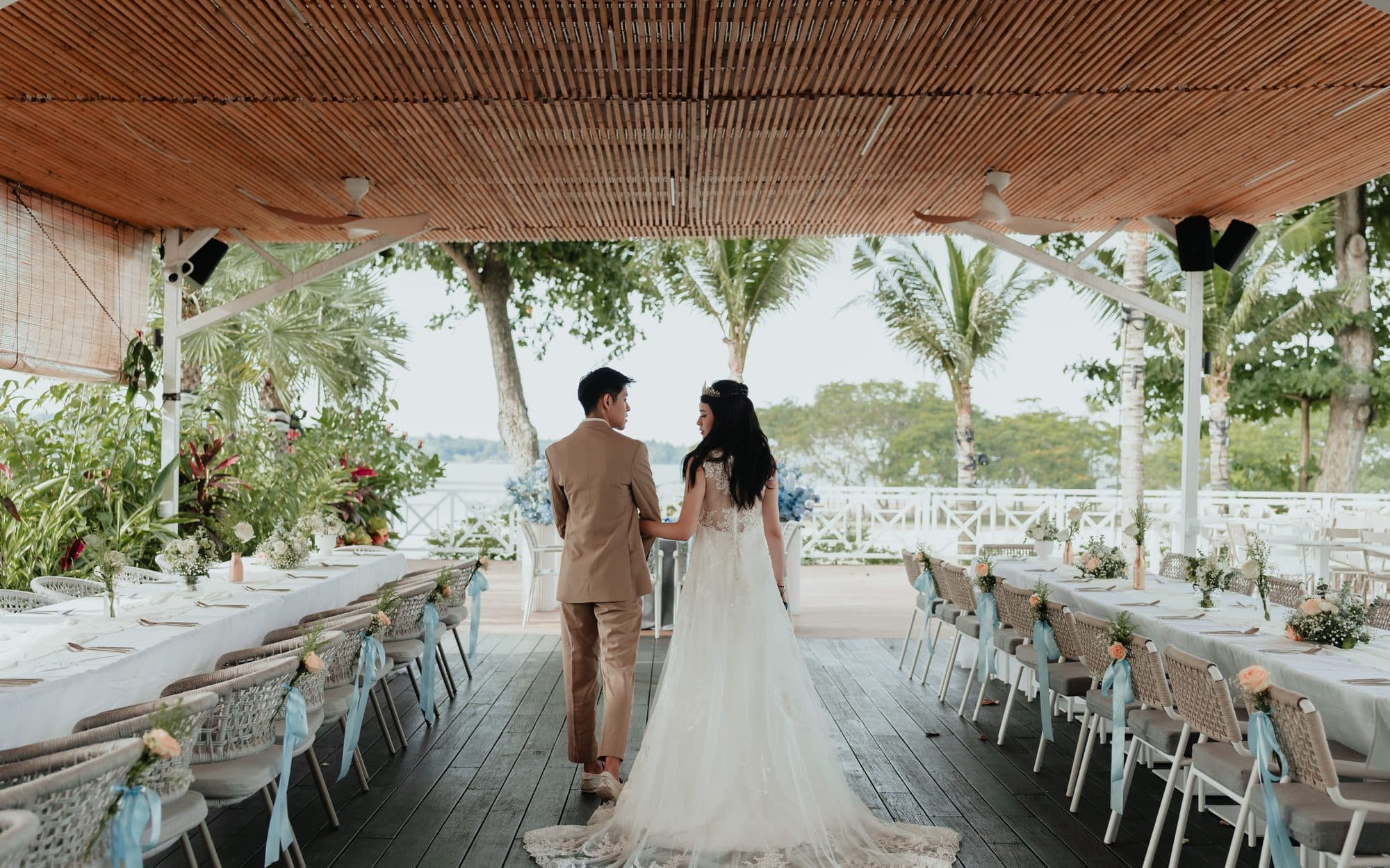 A couple walking between the long tables under the pavilion at Canopy Changi Village, the water beyond