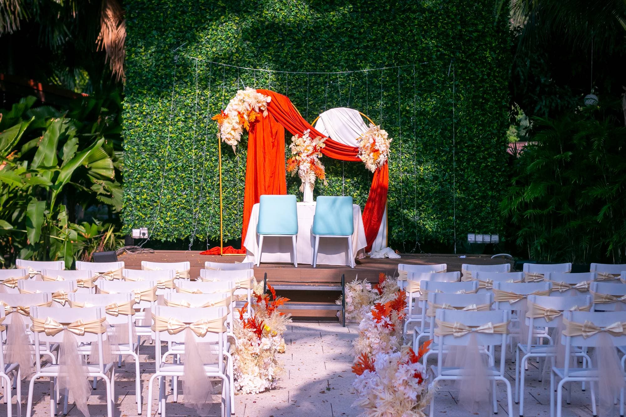 A ceremony arch draped in rust silk against a green hedge, white chairs in rows before it