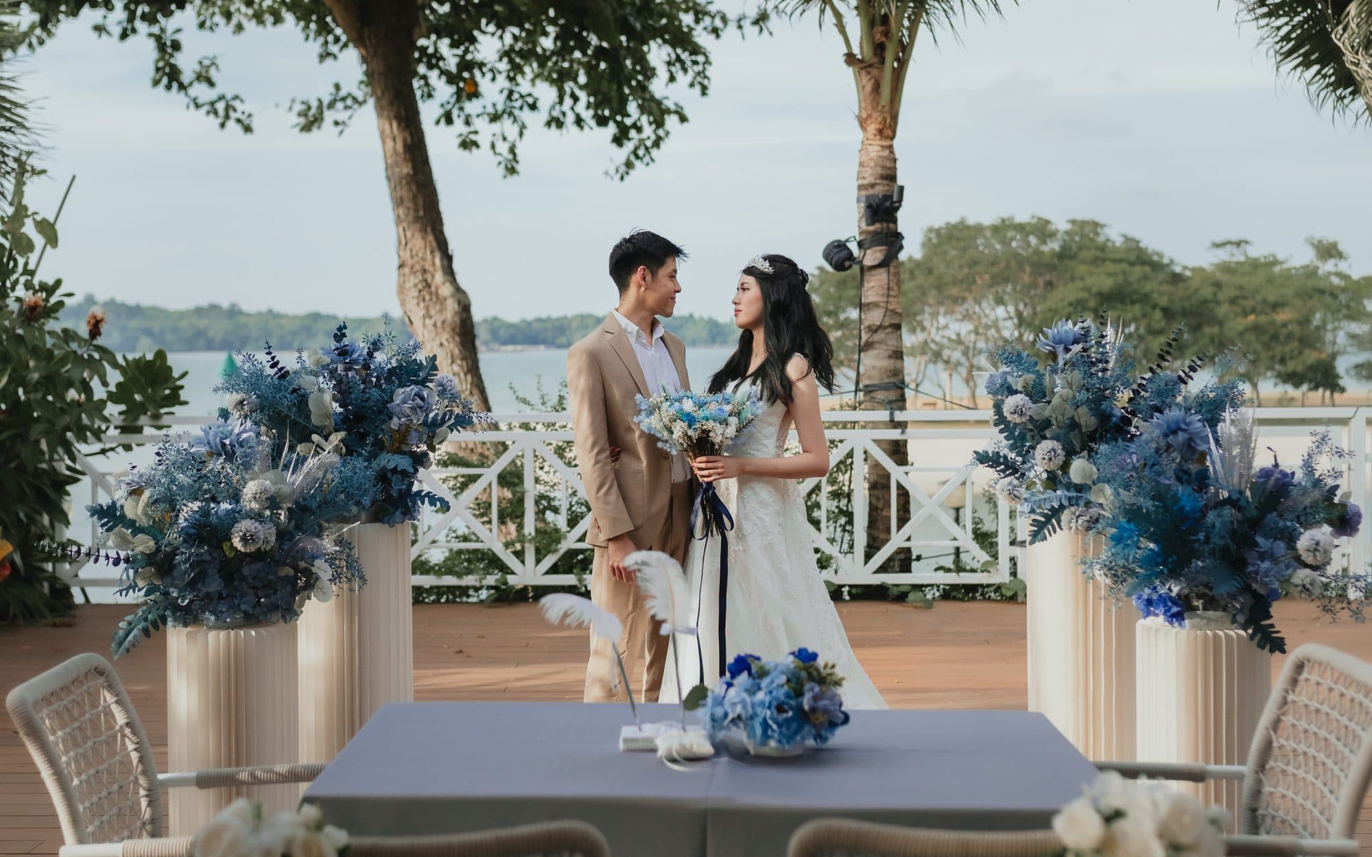 A couple at the ceremony table on the deck at Canopy Changi Village, blue florals either side and the sea behind