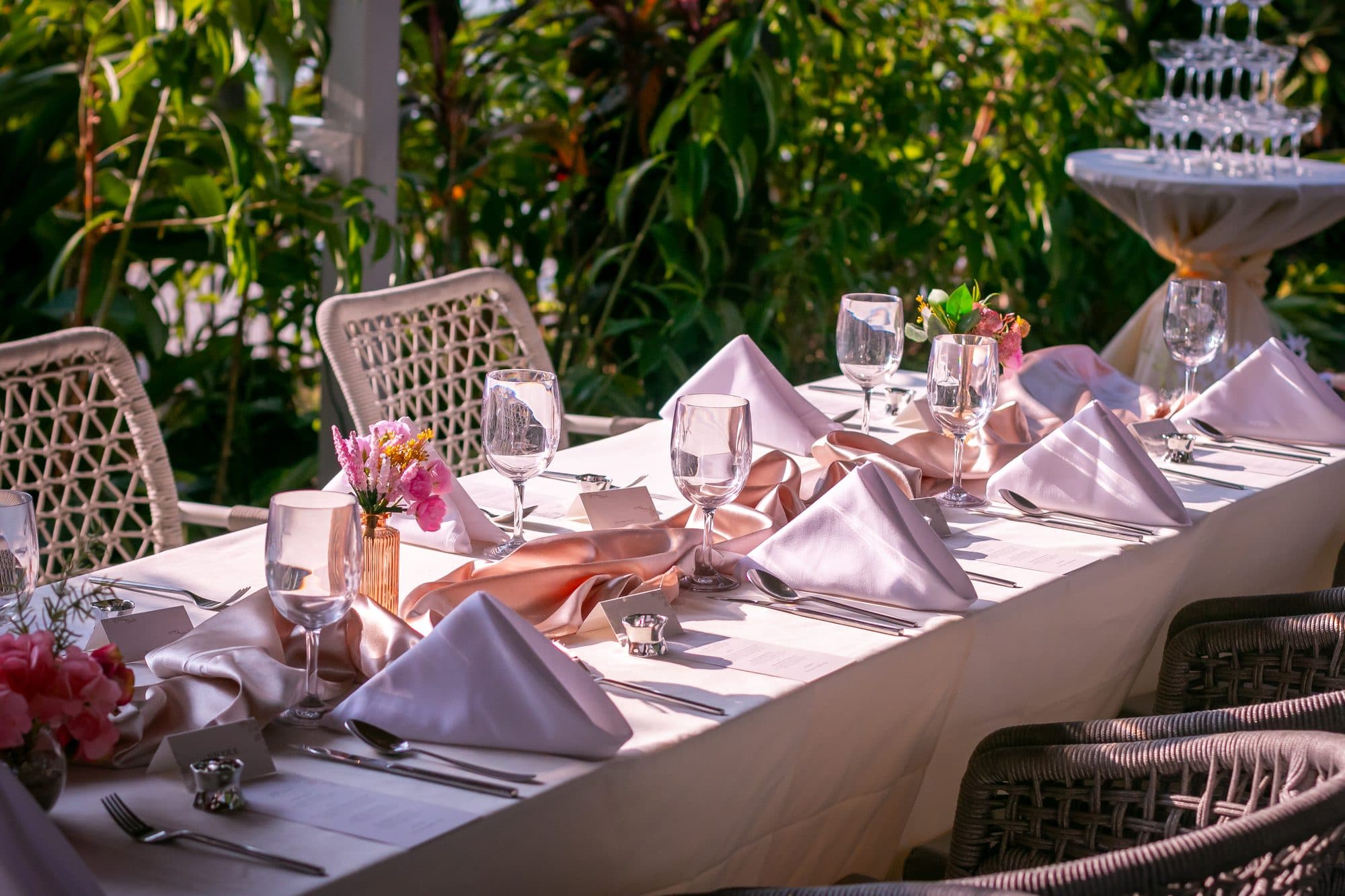 A table laid in blush linen with rattan chairs, a champagne tower behind